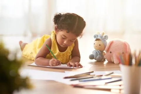Young child in a yellow dress lying on the floor and coloring pages with crayons